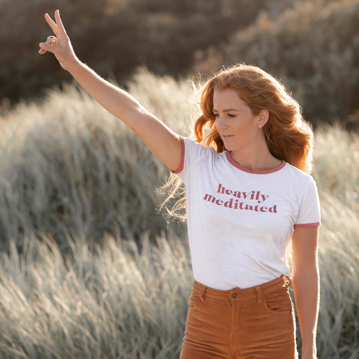 Caitlin Cady standing in front of beach dunes, wearing a Heavily Meditated shirt, her hand raised in a peace sign